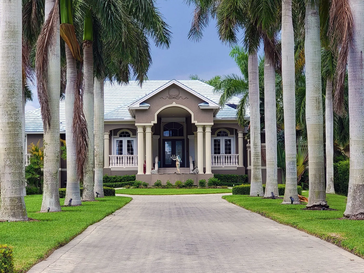 Entrada de residencia exclusiva flanqueada por palmeras reales, con grama pareja y setos definidos a ambos lados del camino